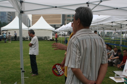 ▲ 19일 오전 7시경 서울광장 중앙 내 분향소 설치가 한창인 가운데 추모객으로 보이는 한 시민이 지난해 광우병 파동 때 자주 사용됐던 '이명박 STOP'이라고 쓰여진 플라스틱 부채를 손에 들고&nbsp;또 다른&nbsp;시민과 이야기를 나누고 있다. ⓒ 뉴데일리