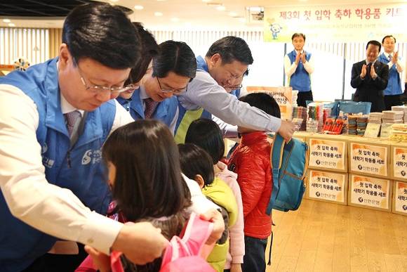 ▲ 신한은행 임원 및 본부장들이 첫 입학 축하 학용품 키트 제작 행사에서 아이들에게 직접 가방을 메어주고 있는 모습.ⓒ 신한은행