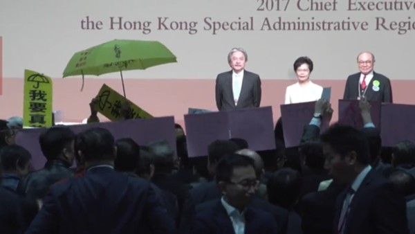▲ 홍콩 행정장관 선거  결과 발표 직후 선거인단에게 인사하는 후보자들. 왼쪽부터 존 창 전 재무사장, 캐리 람 당선인, 우쿽훙 전 고등법원판사. 반중시위대가 선거결과에 항의하여 우산시위 상징물인 노란우산과 '我要真普選'(나는 진정한 직선제를 원한다) 현수막을 들고 있다. (홍콩 明報 사진)