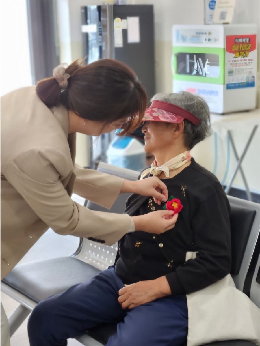 ▲ 제주시서부보건소 직원이 지역 어르신에게 손뜨개로 만든 동백 브로치를 달아드리고 있다. ⓒ제주시 제공